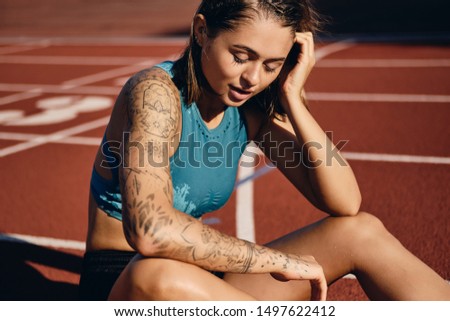 Pensive wet athlete girl with tattooed hand in sportswear resting after running on city stadium