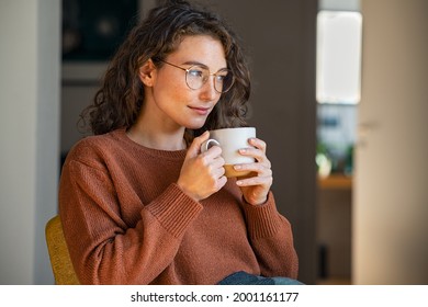 Pensive woman drinking hot coffee at home. Thoughtful young woman drinking a cup of tea while thinking. Pretty contemplative girl with sweater relaxing at home while drinking purifying herbal tea. - Shutterstock ID 2001161177