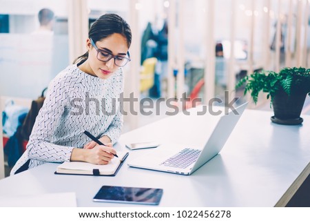 Pensive woman in eyewear planning working schedule writing in notebook while sitting at working place with laptop computer,female administrative manager making notes of information browsed on netbook