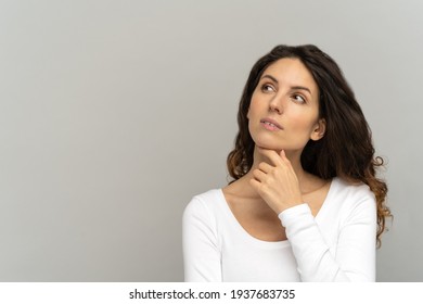 Pensive woman keeps finger on chin, concentrated and ponders, wears white T-shirt with long sleeves, look up at blank copy space. Girl thinking over sale offer making choice, looking aside. Mock up.
