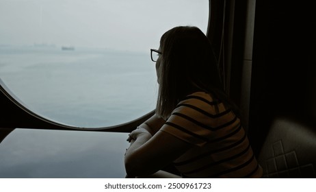 A pensive woman looks out a cruise ship window at the sea reflecting on her journey - Powered by Shutterstock