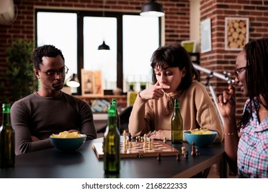 Pensive woman sitting at table while thinking about next chess move. Young smart looking people playing strategy boardgames together while sitting at home in living room.