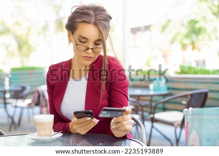 A pensive woman with a smartphone and a credit card sits in a summer cafe. Online shopping concept.