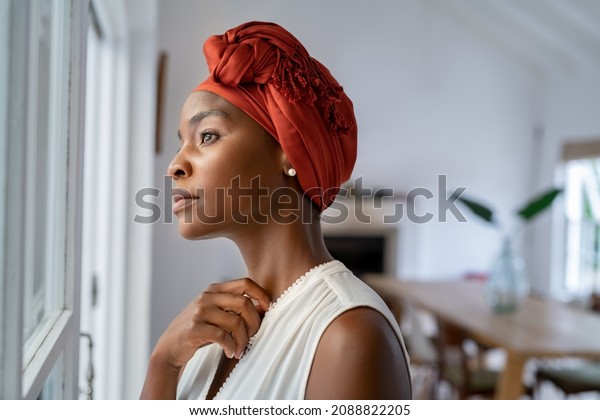 Pensive woman wearing headscarf looking outside\
window in contemplation. Mature black woman wearing a traditional\
turban and thinking near window at home. Worried african mature\
woman with cancer.