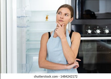 Pensive young adult housewife in apron standing on kitchen near opened fridge with leftover apple at empty shelf, ponder , looking away