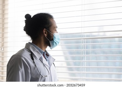 Pensive Young African American Biracial Gp Doctor Therapist In Respirator Facemask Looking In Distance Out Of Window, Thinking Of Problem Solution, Pandemic Situation Or Illness Treatment In Clinic.