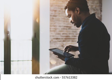 Pensive young African American freelancer touching screen digital tablet while working in new project at home.Black businessman looking for information on Internet.Blurred,color filter,flares