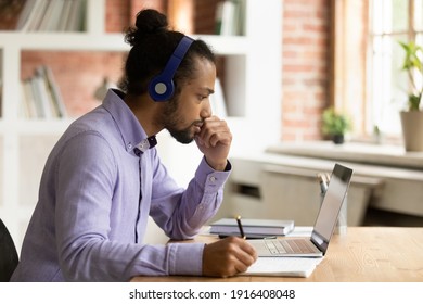 Pensive Young African American Man In Headphones Look At Laptop Screen Thinking Studying Online From Home. Thoughtful Ethnic Male Student In Earphones Take Distant Course On Computer Learning.