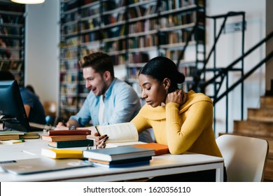 Pensive Young African American Woman In Casual Clothes Reading Book With Interest Sitting At Table Beside Excited Millennial Man Using Computer And Taking Notes In Notepad In Contemporary Library