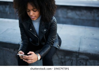 Pensive Young African American Woman With Bouncy Hair In Black Coat Looking For Information On Internet While Chilling On Curb In City