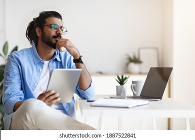 Pensive Young Arab Male Freelancer Using Gigital Tablet And Daydreaming At Home, Handsome Eastern Man Thinking About New Business Ideas, Sitting At Desk Touching Chin And Looking Aside, Free Space
