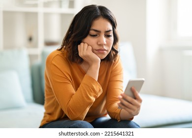 Pensive Young Arab Woman Looking At Smartphone Screen While Sitting On Couch At Home, Upset Millennial Middle Eastern Female Feeling Bored Or Waiting For Important Call Or Sms, Copy Space - Powered by Shutterstock