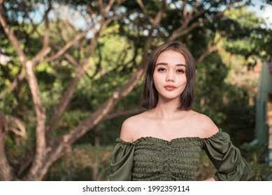 A pensive young asian lady wearing a green off shoulder blouse during a windy day at the park outdoors.