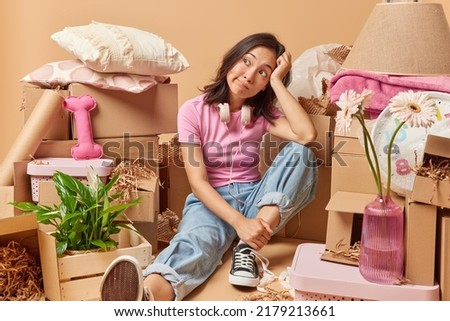 Pensive young Asian woman sits tired on floor thinks about something dressed in casual clothes relocates in new house surrounded by cardboard boxes full of personal belongings going to move in