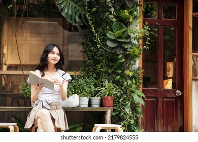 Pensive Young Asian Woman Sitting Outside Cafe And Reading Interesting Book