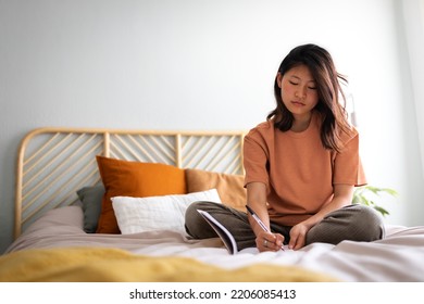 Pensive Young Asian Woman Sitting On Bed With Serious Expression Writing On Journal In Cozy Bedroom. Copy Space.