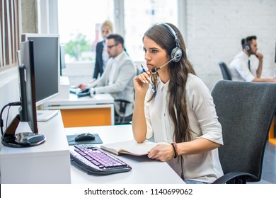 Pensive young attractive business woman with headset holding pen and notepad and looking something on computer screen - Shutterstock ID 1140699062