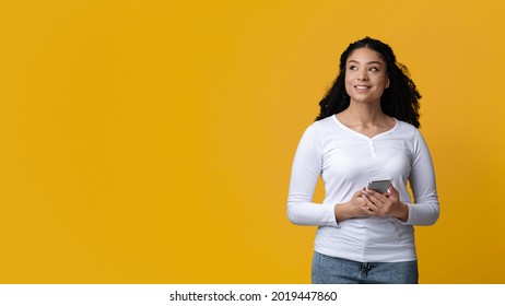 Pensive Young Black Woman Holding Smartphone In Hands And Looking Away, Dreamy African American Female Thinking About Mobile Offer, Standing Isolated Over Yellow Background, Panorama With Copy Space