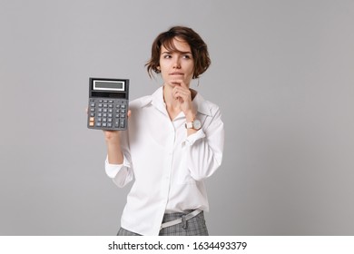Pensive young business woman in white shirt posing isolated on grey wall background in studio. Achievement career wealth business concept. Mock up copy space. Hold calculator put hand prop up on chin - Shutterstock ID 1634493379