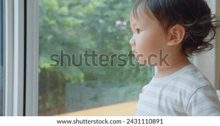 A pensive young child gazes out a window into a lush garden, lost in thought, with soft natural light accentuating a peaceful expression