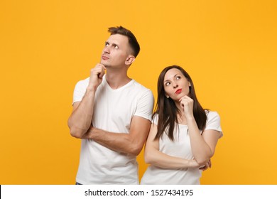 Pensive young couple two friends guy girl in white empty blank t-shirts posing isolated on yellow orange background. People lifestyle concept. Mock up copy space. put hand prop up on chin, looking up