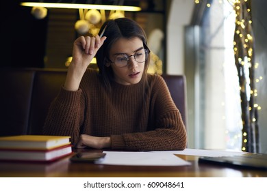 Pensive young female skilled student in glasses and casual outfit checking report correcting mistakes before sending to coach proofreading in coffee shop during break between lessons in university 