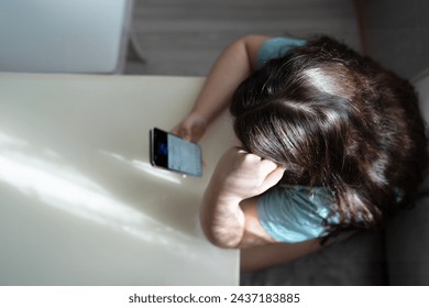 A pensive young girl sits at a table, deeply engrossed in the content on her smartphone. The indoor setting is bathed in natural light filtering through lace curtains, creating a tranquil domestic sce