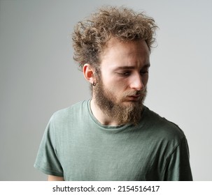 Pensive Young Handsome Bearded Man With Wild Curly Hair, Bright Blue Eyes Looking Down Isolated On White Background. Young Thinking Man In Green T Shirt On White. 