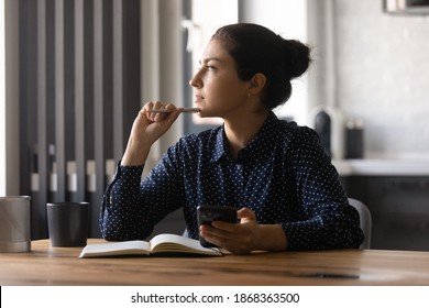 Pensive young hindu female study by desk using mobile internet distracted from making notes create new idea. Thoughtful mixed race woman looking aside of phone screen pondering planning future work