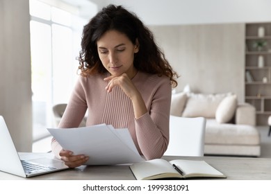 Pensive Young Hispanic Woman Sit At Desk At Home Office Work Online On Computer Read Paperwork Documents. Millennial Latino Female Use Laptop Consider Financial Report Or Paper Correspondence.