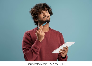 Pensive young Indian man, businessman, freelancer, entrepreneur holding a notepad and dreamily looking up thinking on new business project, writing list-to-do, isolated on blue color background - Powered by Shutterstock