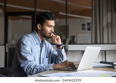 Pensive young Indian manager man working at laptop, sitting at workplace table, looking at display, typing, touching chin, thinking on online content, using computer for job communication - Powered by Shutterstock
