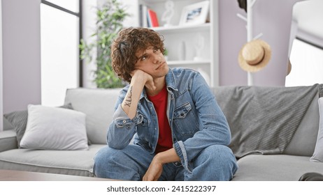 Pensive young man with curly hair, wearing denim, sitting on a couch in a modern living room. - Powered by Shutterstock