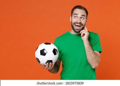 Pensive young man football fan in green t-shirt cheer up support favorite team with soccer ball put hand prop up on chin isolated on orange background studio. People sport leisure lifestyle concept - Shutterstock ID 1815975839