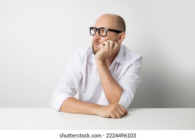 Pensive Young Man Holding Hand Under Chin While Sitting At Table On White Background.