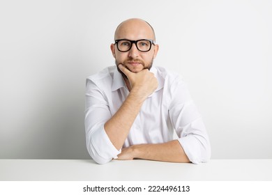 Pensive Young Man Holding Hand Under Chin While Sitting At Table On White Background.