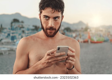 Pensive Young Man with Smartphone on Beach - A young Caucasian man in swimwear holds a smartphone, gazing off-camera with a questioning look, with blurred beach in the background. - Powered by Shutterstock