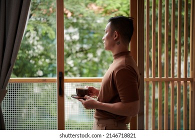 Pensive young man standing on balcony, drinking morning coffee and looking at street - Powered by Shutterstock