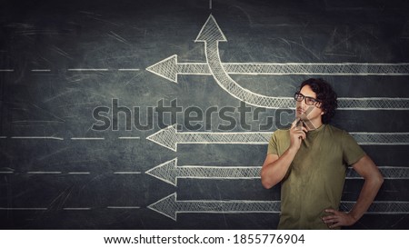 Pensive young man stands over chalkboard background with multiple straight arrows sketches, one of them changes direction bending by upwards trajectory. Different thinking concept, breaking the rules