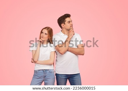 Pensive young man and woman in similar clothes touching chins and thinking, while standing near each other and looking in different directions against pink background