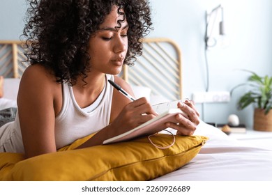 Pensive young multiracial latina woman lying down on bed with serious expression writing on journal in cozy bedroom - Powered by Shutterstock