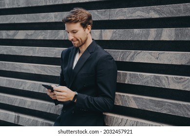 Pensive young office worker in formal stylish suit standing outdoor holding mobile phone in his hands, reading last emails or messages, trying to type response on important urgent correspondence - Shutterstock ID 2032457921