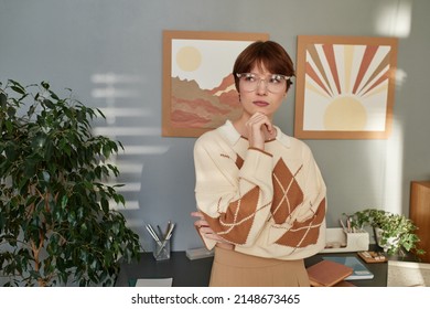 Pensive young redhead teacher in eyeglasses and sweater touching chin and looking aside in modern room
