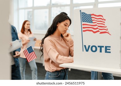 Pensive young woman, Asian voter waiting in line, standing in polling booth with American flag, voting at polling station. Concept of US presidential elections, democracy - Powered by Shutterstock