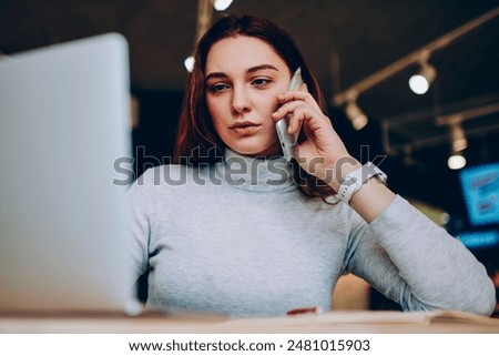 Pensive young woman calling to customers support service updating antivirus program for protection netbook software, concentrated female confirming booking hotel on web page talking on cellular