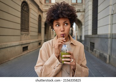 Pensive young woman with curly hair sips fresh vegetable cocktail eats fitness food for weight loss dressed in brown jacket strolls in ancient city thinks about something. Nutrition concept.
