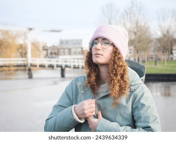 A pensive young woman with curly hair and glasses zips up her teal jacket while standing by a riverside, wearing a pink beanie - Powered by Shutterstock