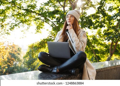 Pensive young woman dressed in autumn coat and hat sitting outdoors, using laptop computer