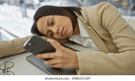 A pensive young woman dressed in business casual attire rests on a table while gazing at her smartphone. - Powered by Shutterstock