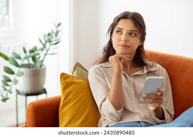 Pensive young woman holding smartphone and looking away while relaxing on couch at home, beautiful female deep in thoughts using mobile phone in domestic interior, thinking and planning something - Powered by Shutterstock